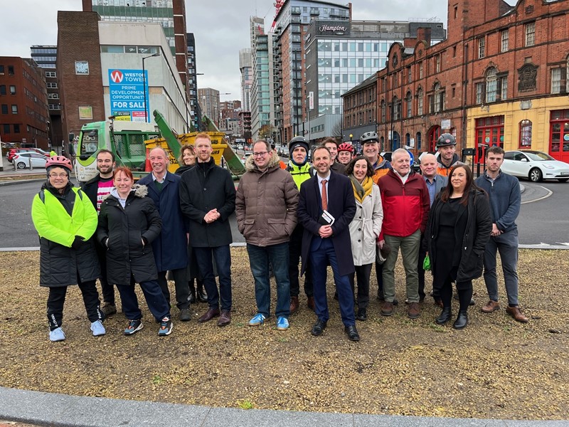 A photo of 18 people standing on a roundabout that has been built in December 2024. The people are invited guests and councillors wearing coats and smiling at the camera.