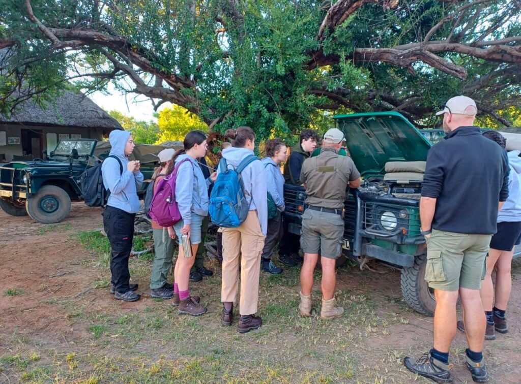 Students and leaders gather around a safari vehicle with the hood open and discussing what they see