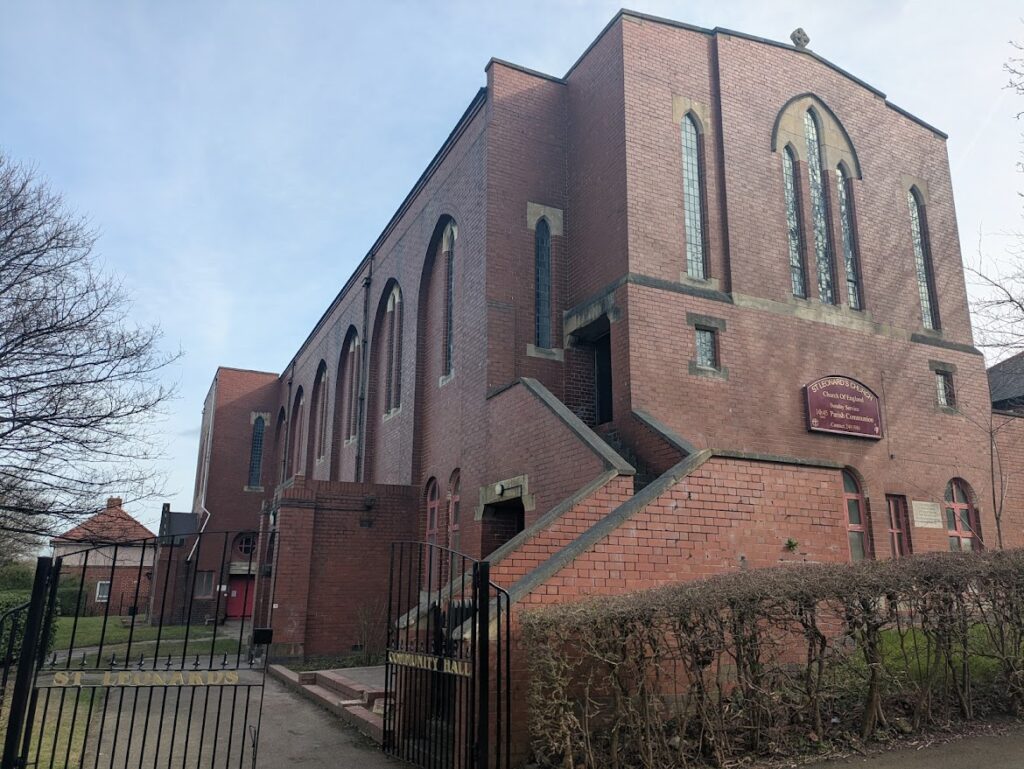 The exterior of the red brick church building, with a placard on the front saying 'St Leonard's Church'. There is a metal gate in the foreground which is the entrance to the nursery.