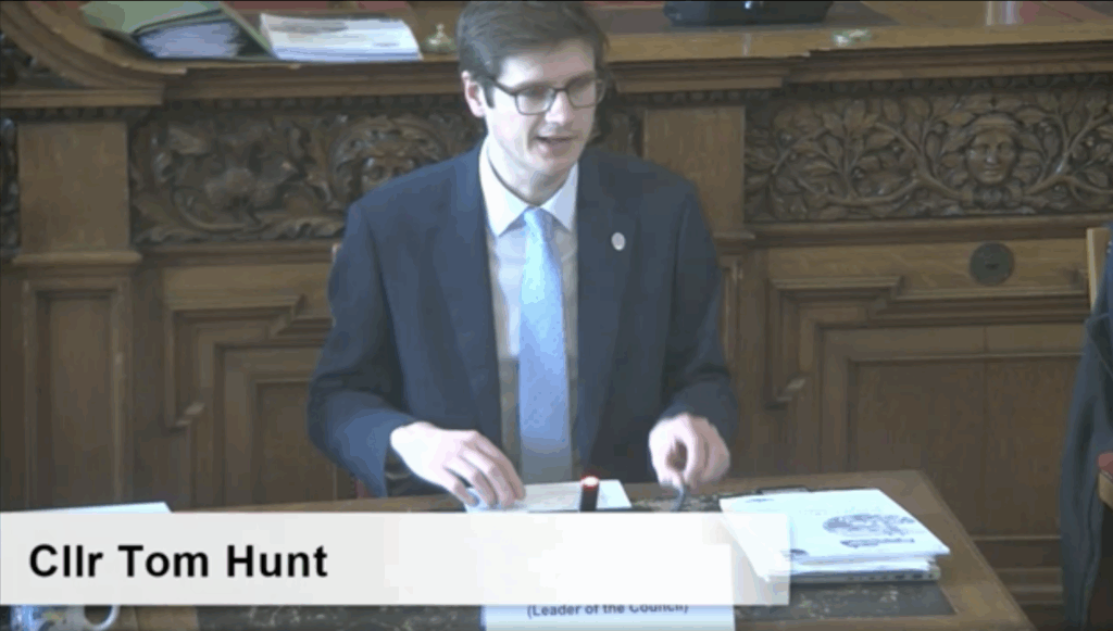 A man wearing glasses, a blue blazer and a white shirt is speaking in court. He is sat on a red chair and has sheets of paper stacked in front of him. An on-screen graphic displays his name "CLLR TOM HUNT".