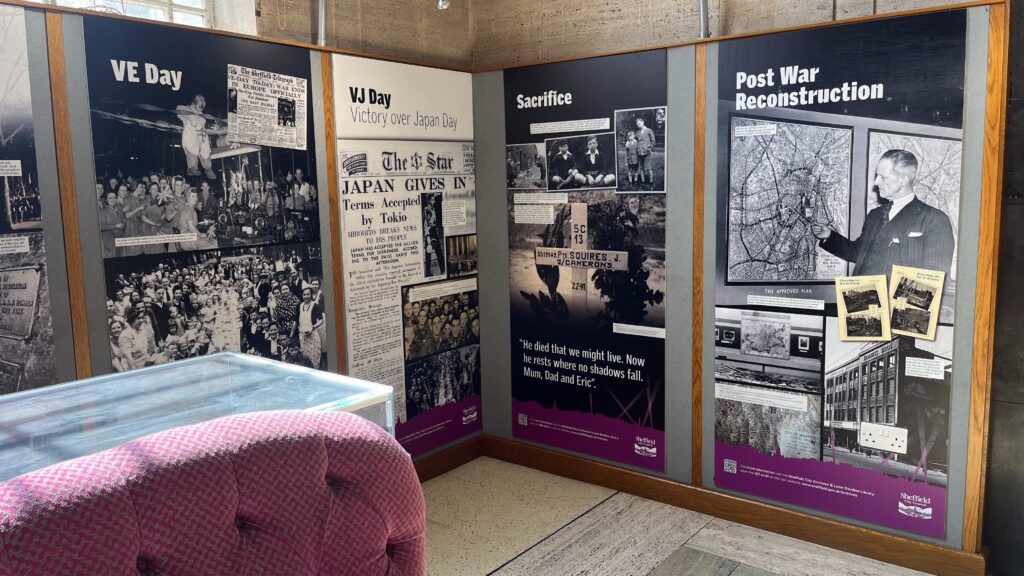 A VE Day exhibition in Sheffield's Central Library. In the corner there are four informational panels about the end of World War II, including VE Day, VJ Day, Sacrifice, and Post War Reconstruction. A glass display case and a pink sofa are in the foreground.