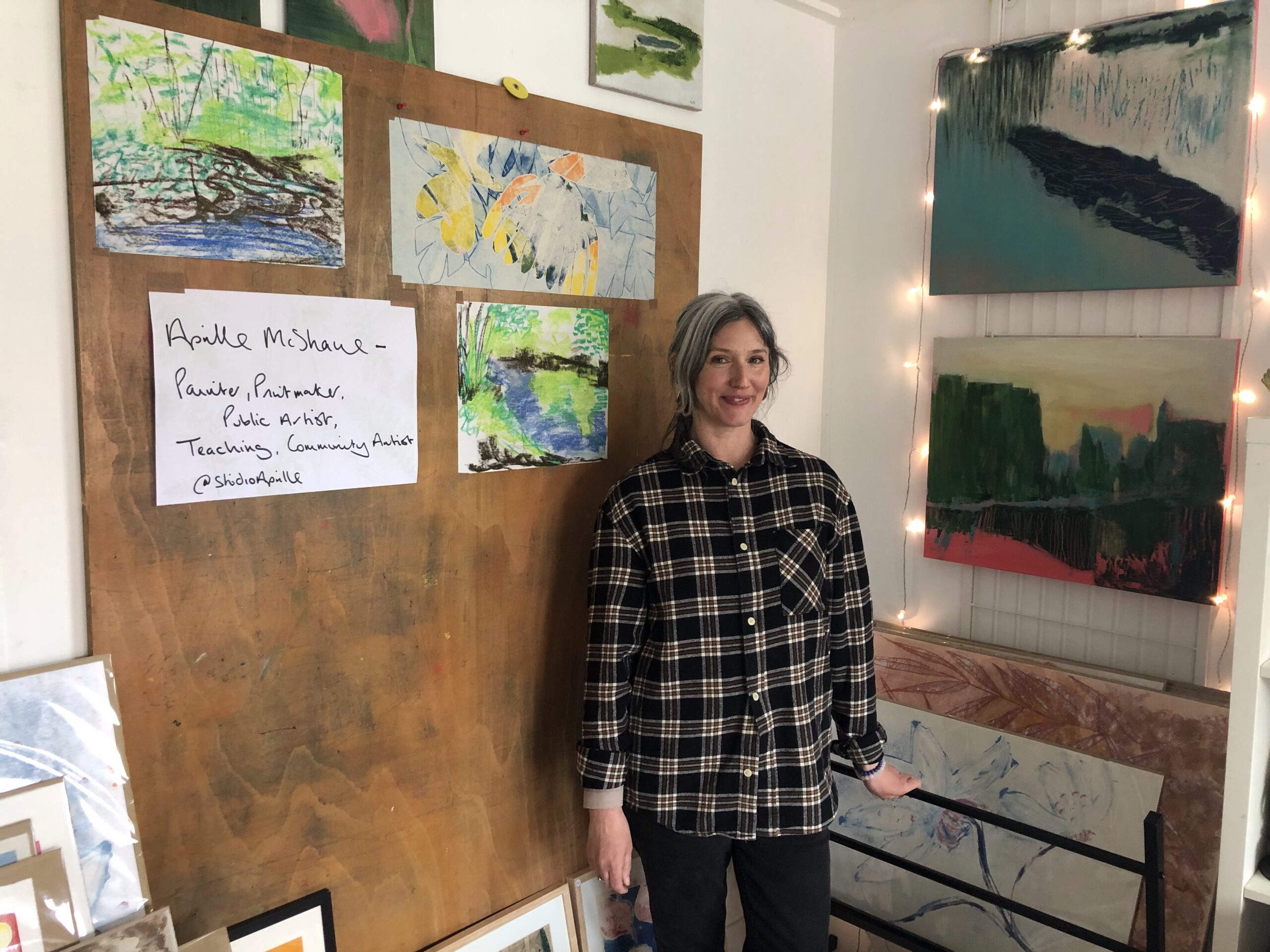 Artist stands in front of wooden board with art pieces and fairy lights