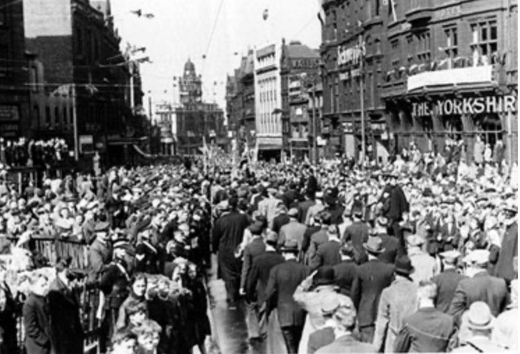A black and white photograph shows a large crowd of people filling a Fargate, Sheffield for a VE Day Victory Parade. Many people appear to be looking towards something in the distance or celebrating. Flags hang from some of the buildings. A sign that reads "THE YORKSHIRE" is visible on one of the buildings to the right.
