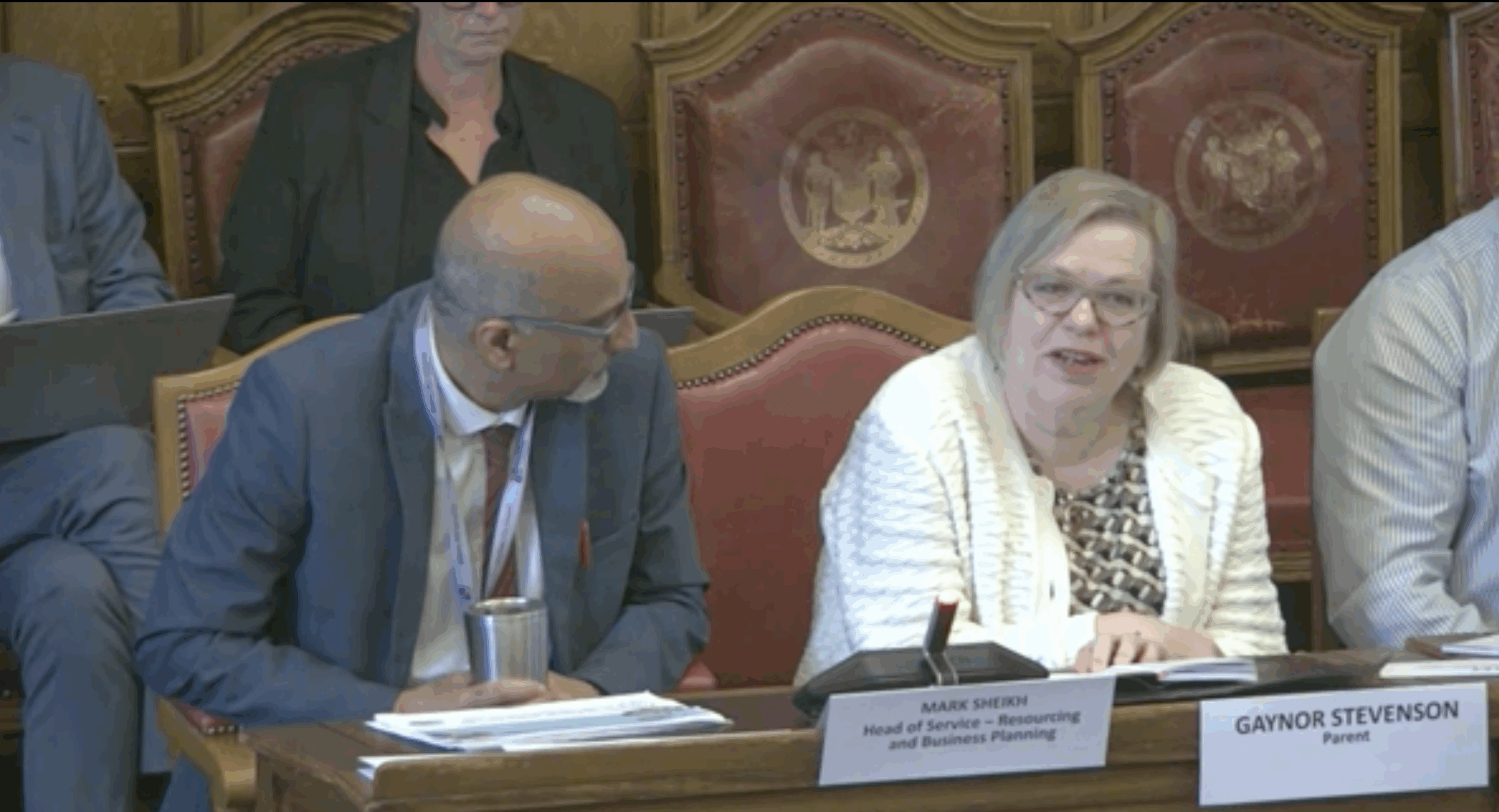 A woman with short grey hair and glasses speaks at a table with name cards reading "MARK SHEIKH, Head of Service - Resourcing and Business Planning" and "GAYNOR STEVENSON, Parent". Mark, man with a bald head and glasses, listens to Gaynor speak.