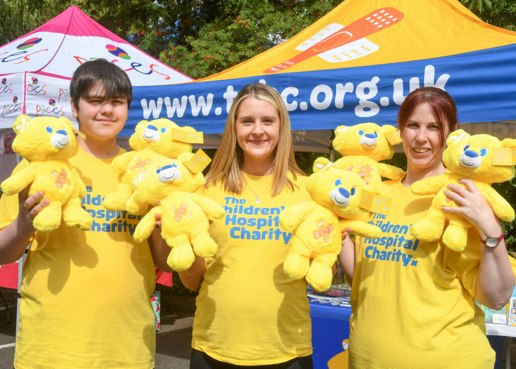 Three individuals, one male and two females, are standing outdoors in front of colorful tents. They are all wearing yellow t-shirts with "Sheffield's Children's Hospital Charity" logo and are holding multiple yellow teddy bears.