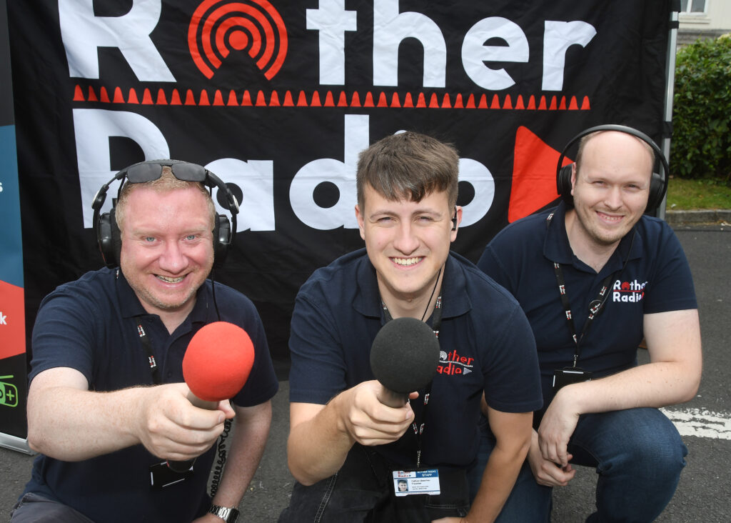 Three men wearing dark blue polo shirts and headphones are holding microphones towards the viewer and smiling. A black banner with "Rother Radio" written in white and red is behind them.