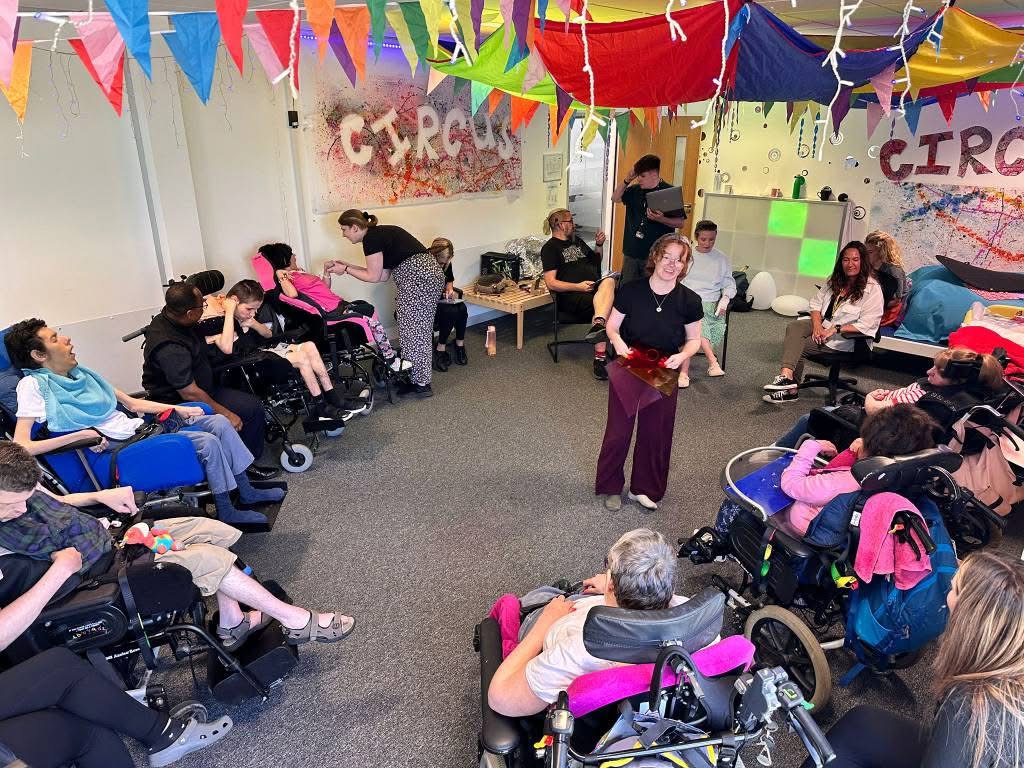 A high-angle, wide shot shows a group of people, many in wheelchairs, gathered in a brightly decorated room, possibly for an activity or performance. Colorful triangular flags and pieces of fabric are strung across the ceiling, and the walls feature vibrant, abstract paint splatters with the word "CIRCUS" written on them in two places.
In the center, a woman with curly reddish-brown hair, wearing a black shirt and dark red pants, stands facing the group, holding a red object. She appears to be leading an activity or presenting something.
Around her, people are seated in a semi-circle, many in various types of wheelchairs. On the left side of the frame, several individuals are visible, some looking towards the center. On the right, more individuals are seated, including some adults and what appear to be younger people.
In the background, other adults are visible standing or seated, some observing the activity. One person in a dark blue uniform is visible near a counter in the far back right. The floor is carpeted in grey, and there are various pieces of equipment and personal belongings scattered around. The atmosphere appears lively and engaging.