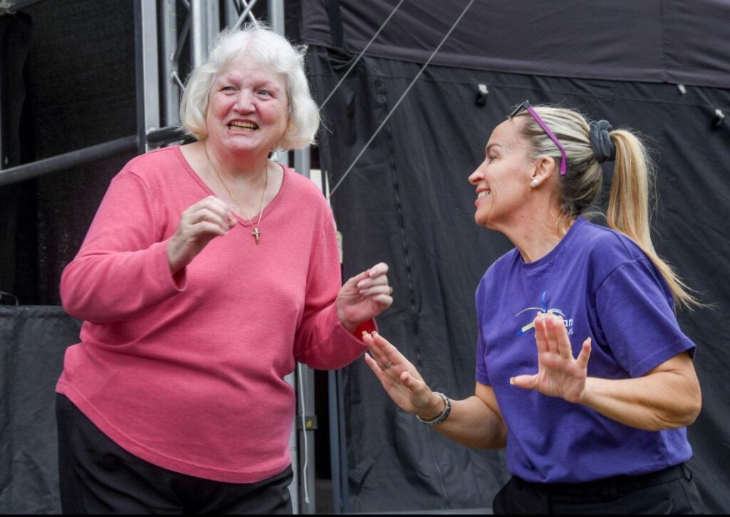 A smiling, fair-skinned woman with short, white hair, wearing a pink long-sleeved shirt and a gold cross necklace, holds her hands up as if in surprise or excitement. To her right, a fair-skinned woman with blonde hair pulled back in a ponytail, wearing a purple t-shirt, laughs and gestures with her hands. Both women appear to be interacting playfully. In the background, there's a dark, possibly black, structure, perhaps a tent or stage backdrop.